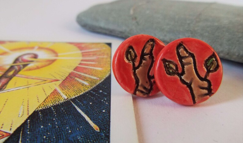 May include: A pair of red ceramic earrings with a hand and leaves design. The hand is brown and the leaves are green.