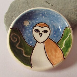 May include: A small ceramic dish with a hand-painted design of a barn owl in a night sky with a full moon. The owl is white with brown markings and is surrounded by green hills.