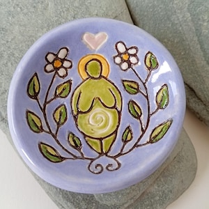 May include: A small, round, ceramic dish with a light purple background. The dish features a green figure with a yellow halo, a pink heart, and white flowers with yellow centers. Dark brown stems and green leaves surround the figure.