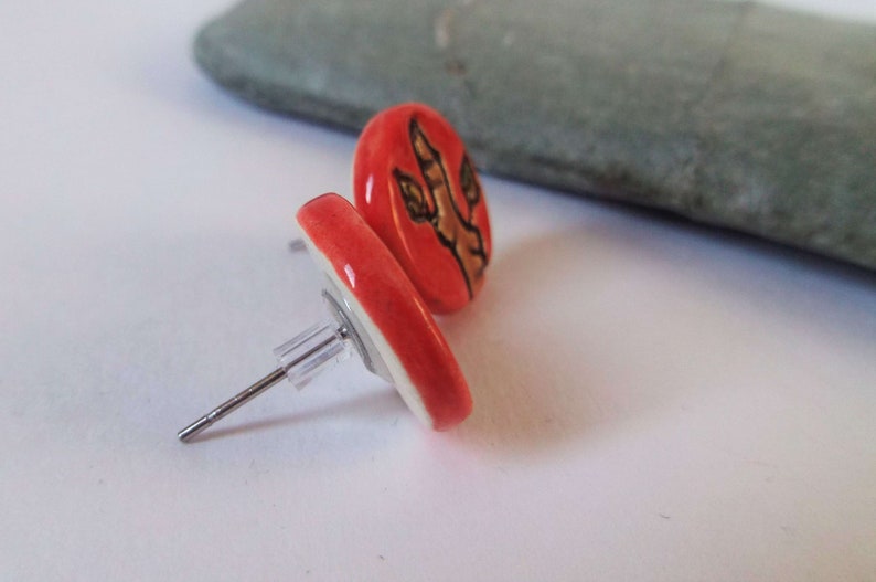 May include: A pair of red ceramic stud earrings with a brown dinosaur footprint design.