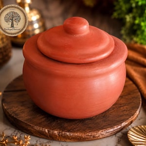 May include: A round terracotta clay pot with a matching lid, featuring a smooth, matte, reddish-brown finish. The pot has a wide, curved body and a simple knob handle on the centre of the lid. A circular logo with a tree illustration and the text KarthigaEthnicart is visible in the upper left corner.