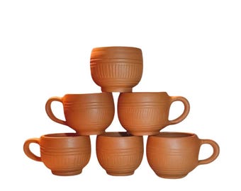 May include: A set of six terracotta-colored clay cups arranged in a pyramid formation. Each cup features a rounded body with a handle and a series of horizontal incised lines and vertical textured patterns encircling the exterior surface.