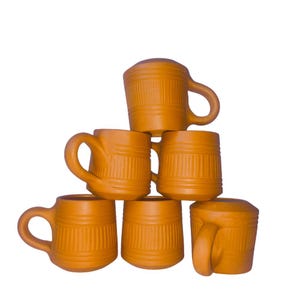 May include: A stack of six terracotta-colored mugs with rounded handles. Each mug has vertical ribbed detailing around the body. The mugs are arranged in a pyramid shape against a white background.
