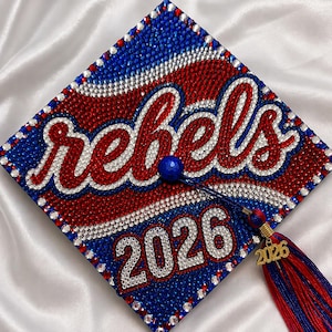 May include: A graduation cap decorated with red, white, and blue rhinestones. The word "rebels" is spelled out in red rhinestones, and the year "2026" is displayed. A tassel with a gold charm that reads "2026" hangs from the cap.