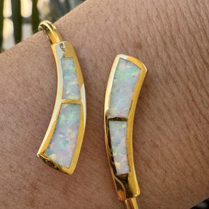 May include: A gold-toned cuff bracelet featuring two curved, open-ended terminals. Each terminal is inlaid with two rectangular segments of iridescent white opal, displaying flashes of green, blue, and pink light against a polished metal surface.