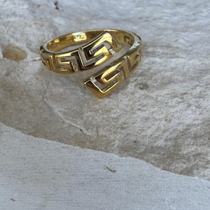 May include: A gold-colored ring with a Greek key design. The ring has an open, wrap-around style. The ring is on a light-colored, textured surface.