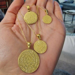 Può includere: Collane con ciondoli dorati con intricati disegni circolari. Le collane variano di dimensioni, con il ciondolo più grande con motivi dettagliati. Le catene sono delicate e i ciondoli sono tenuti in una mano, mostrando il design e il colore dei gioielli.