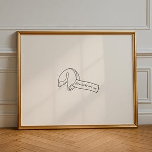 May include: A framed print featuring a minimalist line drawing of a fortune cookie. The fortune reads "How lucky are we" in a handwritten style. The artwork is set against a cream background, framed in gold, and displayed on a wooden floor.