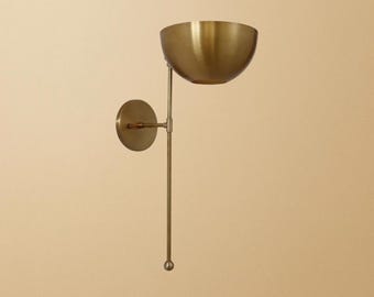 May include: A brass wall sconce with a bowl-shaped shade. The sconce has a circular wall mount and a long, slender arm that supports the shade. The shade is a warm gold color, and the overall design is minimalist and elegant.