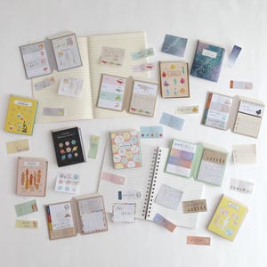May include: An assortment of stationery items, including notebooks, sticky notes, and small booklets, are arranged on a white surface. The items feature various designs, such as dinosaurs, scientific diagrams, and colorful patterns. Some notebooks are open, revealing lined pages.