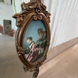 May include: An ornate, gold-colored mirror with an oval-shaped painting of a couple in a carriage. The mirror has a decorative frame with floral and scroll details. The painting features a woman in a pink dress and a man in a red coat.