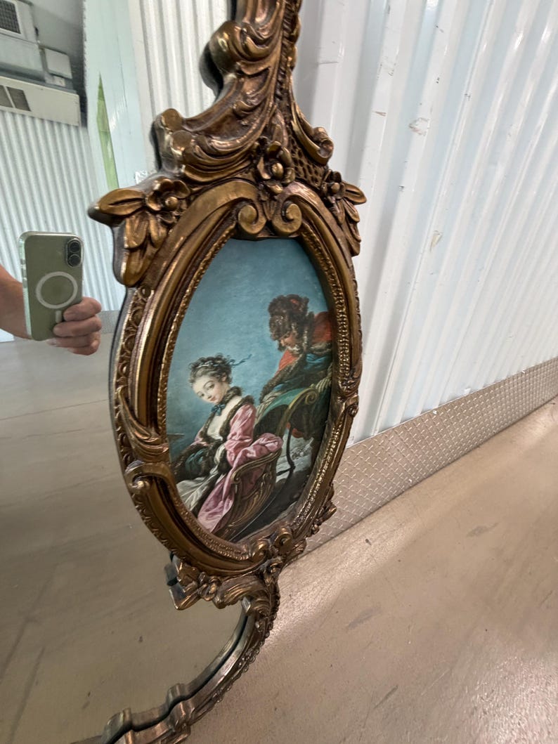 May include: An ornate, gold-colored mirror with an oval-shaped painting of a couple in a carriage. The mirror has a decorative frame with floral and scroll details. The painting features a woman in a pink dress and a man in a red coat.