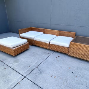 May include: A light brown wooden sectional sofa with white cushions and a matching ottoman. The sofa has a low profile and a modern design. The ottoman is square and has a white cushion. The sofa and ottoman are made of wood.