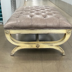 May include: A tufted, light brown leather ottoman with a distressed white painted wooden frame. The ottoman has a curved base with ornate details.