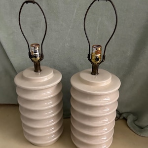 Könnte beinhalten: Paar cremefarbene Keramik-Tischlampen. Jede Lampe hat ein gestapeltes, geriffeltes Design am Sockel und einen dunkelbraunen Lampenschirmhalter aus Metall. Die Lampen haben eine Vintage-Ästhetik und eignen sich für ein Wohn- oder Schlafzimmer.