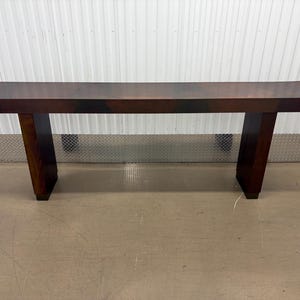 May include: A long, rectangular console table featuring a dark brown wood grain finish. The table has a flat, smooth top surface supported by two thick, solid vertical legs positioned at each end, creating a clean, linear, and structural design.