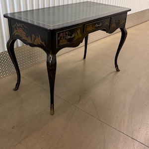 May include: A black writing desk with curved legs and a green leather top. The desk features gold-colored painted landscape illustrations on the front drawers and sides, with gold accents on the feet and edges of the rectangular surface.
