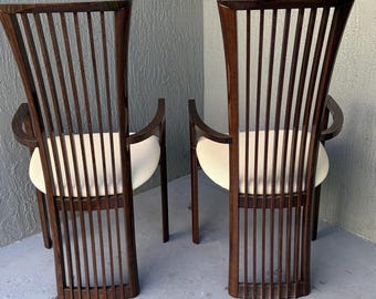 May include: Two wooden chairs with a dark brown finish. The chairs have a slatted back design and curved armrests. The seats are upholstered in a cream-colored fabric. The chairs are placed against a textured gray wall.