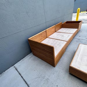 May include: A wooden outdoor sofa and matching coffee table set. The sofa has a light-colored cushion and a slatted wood design. The coffee table has a similar design. The set is made of light brown wood.