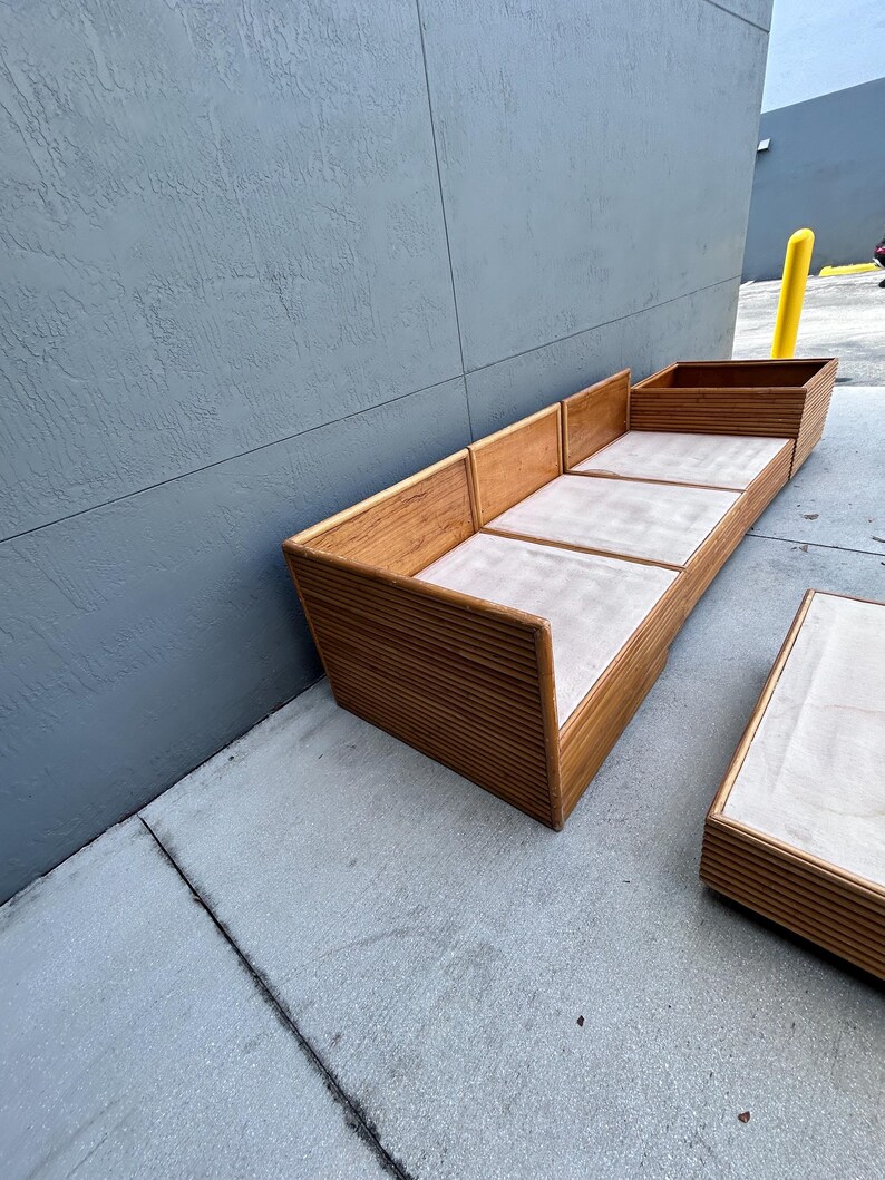 May include: A wooden outdoor sofa and matching coffee table set. The sofa has a light-colored cushion and a slatted wood design. The coffee table has a similar design. The set is made of light brown wood.