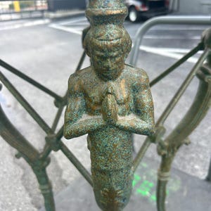 K&ouml;nnte beinhalten: Ein gr&uuml;ner Metalltischfu&szlig; mit einer dekorativen Figur einer Person, die ihre H&auml;nde zum Gebet gefaltet hat. Die Figur steht und hat eine patinierte Oberfl&auml;che.