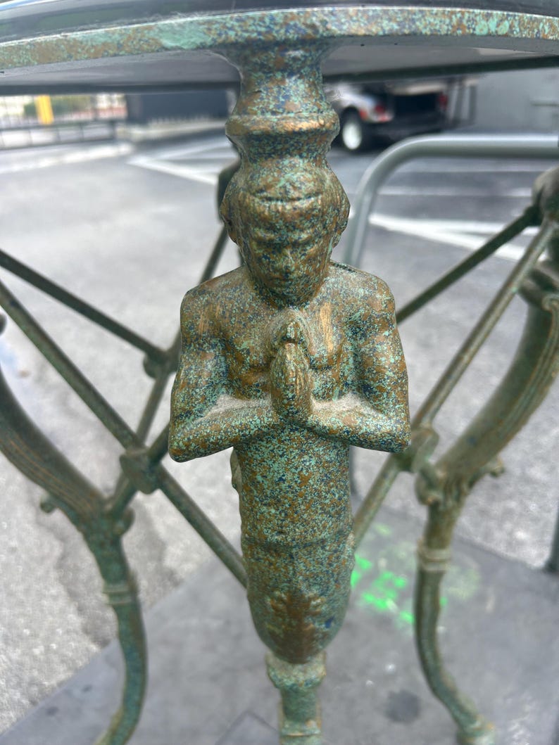K&ouml;nnte beinhalten: Ein gr&uuml;ner Metalltischfu&szlig; mit einer dekorativen Figur einer Person, die ihre H&auml;nde zum Gebet gefaltet hat. Die Figur steht und hat eine patinierte Oberfl&auml;che.