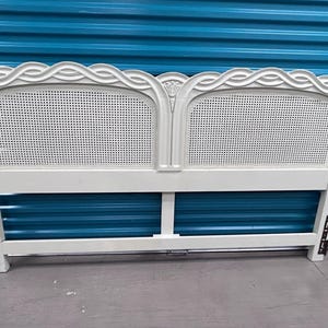 May include: Off-white headboard with a woven cane center and decorative scrollwork. The headboard has a rectangular shape with a wavy design at the top. The headboard is made of wood and has metal brackets.