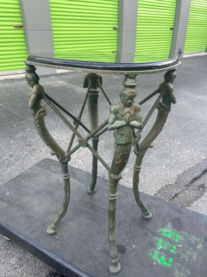K&ouml;nnte beinhalten: Ein gr&uuml;ner Metall-Beistelltisch mit einer schwarzen Glasplatte. Der Tisch hat vier Beine, jedes mit einer Skulptur einer stehenden Person. Der Tisch steht auf einer schwarzen Oberfl&auml;che.