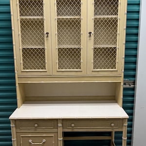 May include: A light yellow desk and hutch combination with a bamboo-style frame. The hutch has three glass-fronted doors with a diamond-patterned mesh. The desk has drawers and a white tabletop. The number 142 is visible on the top.