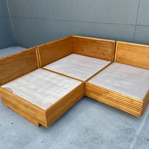 May include: A light brown, modular sectional sofa with a natural wood frame and light-colored cushions. The sofa is composed of three connected sections, forming an L-shape. The frame has a slatted design on the sides.