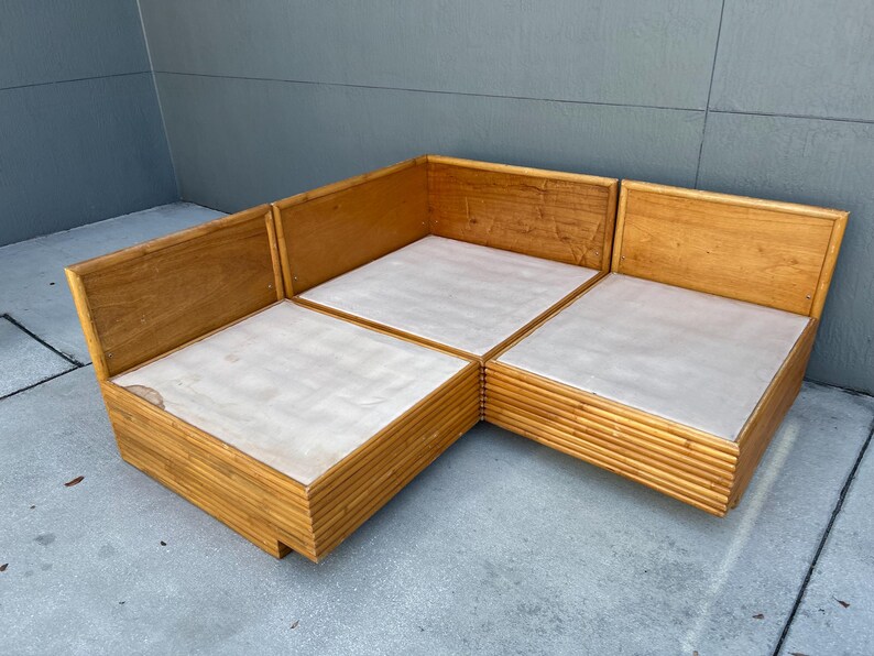 May include: A light brown, modular sectional sofa with a natural wood frame and light-colored cushions. The sofa is composed of three connected sections, forming an L-shape. The frame has a slatted design on the sides.