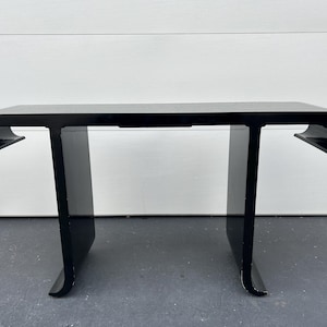May include: A modern black console table with a glossy finish. The table features a rectangular top with curved edges and two T-shaped legs. The table is made of a solid material and is suitable for a living room or entryway.