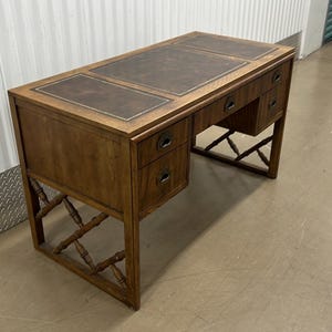 May include: A wooden desk with a brown leather top and multiple drawers. The desk has a rectangular shape with a dark brown leather insert on the top surface. The legs have a decorative lattice design.