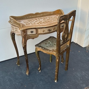May include: An antique vanity set with a matching chair. The set features a small desk with two drawers and a scalloped edge, and a chair with a decorative back. Both pieces are painted in gold and cream with floral accents.