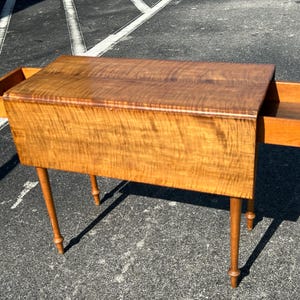 May include: A drop-leaf table with a light wood finish and two drawers. The table has turned legs and a simple design.