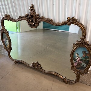 May include: Ornate, antique mirror with a gold-colored, scrolling frame. The mirror features two oval-shaped paintings on each side, depicting figures in period clothing. The mirror is a decorative piece, likely for a living room or bedroom.