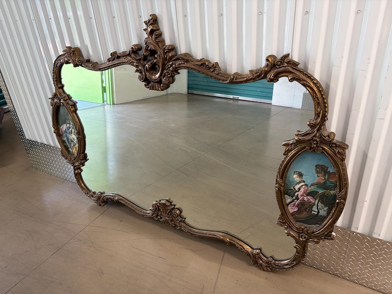 May include: Ornate, antique mirror with a gold-colored, scrolling frame. The mirror features two oval-shaped paintings on each side, depicting figures in period clothing. The mirror is a decorative piece, likely for a living room or bedroom.