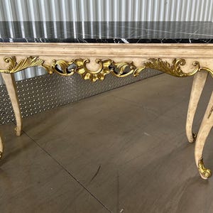 May include: An antique console table with a black marble top and ornate gold and cream-colored accents. The table features curved legs and decorative gold leaf detailing along the apron. A classic piece of furniture.