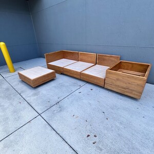 May include: Outdoor wooden sectional sofa and matching square coffee table. The sofa has a light wood finish with a slatted design. The coffee table has a similar design and finish. The furniture is placed on a concrete surface.