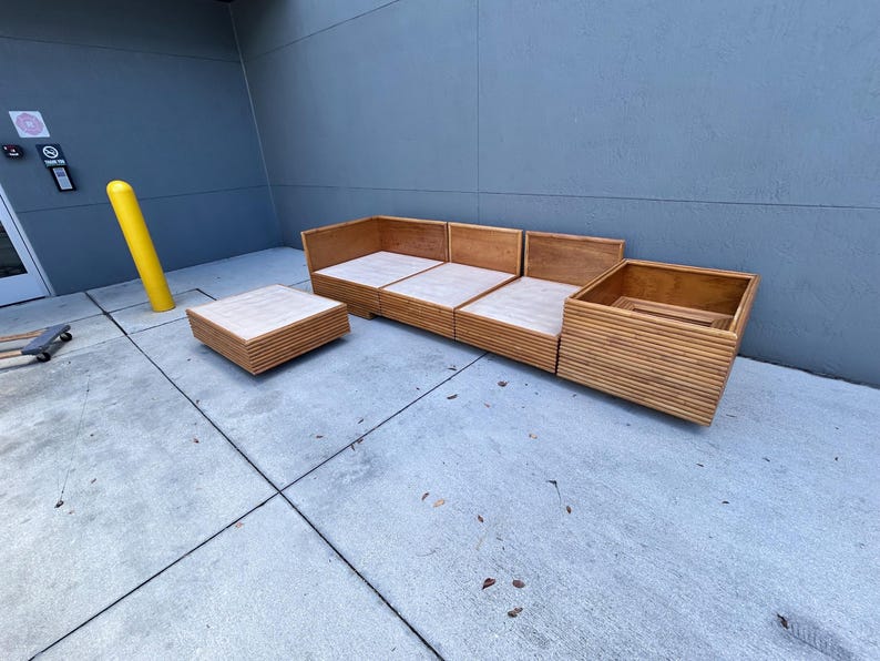 May include: Outdoor wooden sectional sofa and matching square coffee table. The sofa has a light wood finish with a slatted design. The coffee table has a similar design and finish. The furniture is placed on a concrete surface.
