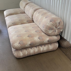 May include: A beige sofa with a patterned design. The sofa has a rectangular base with three seat cushions and four cylindrical back cushions. The fabric has a repeating pattern of feather-like shapes in shades of beige, pink, and gray.