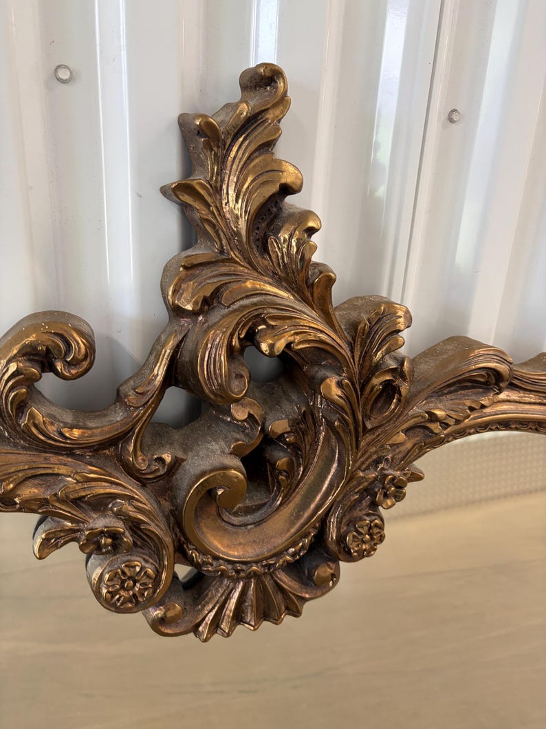 May include: Ornate, gold-colored decorative mirror frame with a detailed, scrolling leaf and floral design. The frame has a vintage aesthetic, with a slightly distressed finish. The design is symmetrical and features intricate carvings.