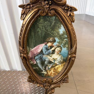 May include: Ornate gold-colored mirror with an oval-shaped frame. The mirror features a painted scene of a couple in a garden setting. The frame has floral and scrollwork details, adding to its antique aesthetic.
