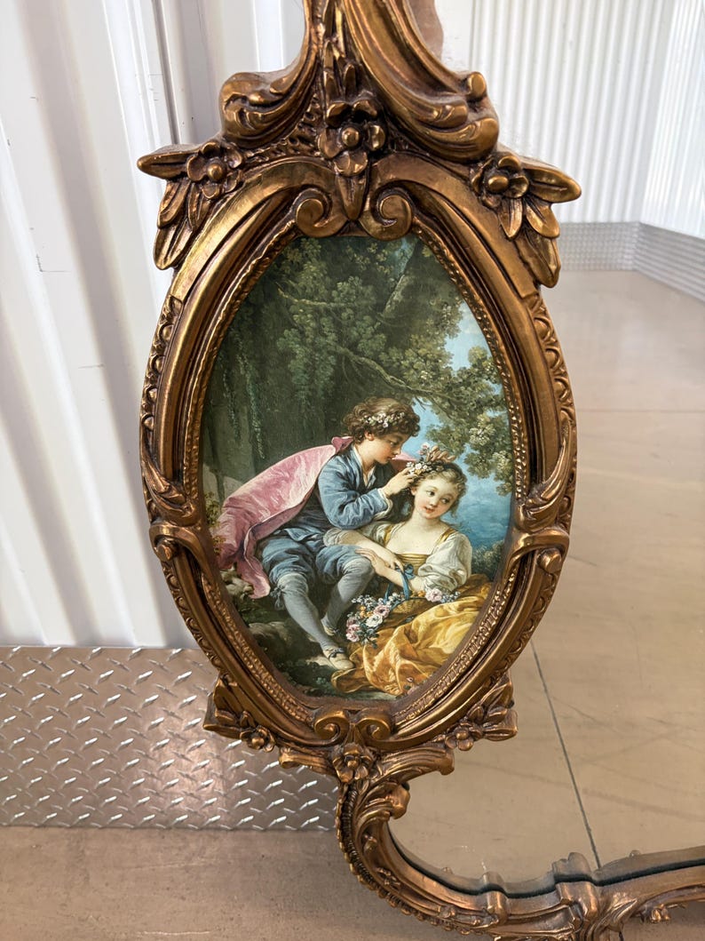 May include: Ornate gold-colored mirror with an oval-shaped frame. The mirror features a painted scene of a couple in a garden setting. The frame has floral and scrollwork details, adding to its antique aesthetic.