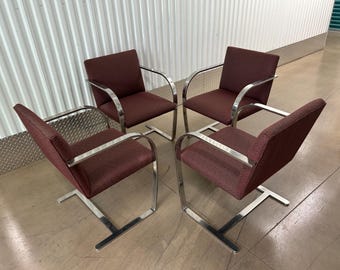 May include: Four burgundy armchairs with chrome frames. The chairs have a low back and a square seat. The armrests are curved and extend from the back to the front of the seat. The legs are a T-shape.