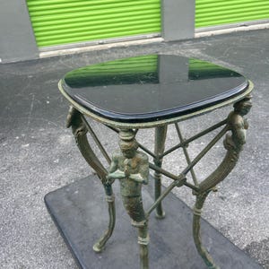 K&ouml;nnte beinhalten: Ein Beistelltisch mit einer schwarzen Marmorplatte und einem gr&uuml;nen Metallgestell mit dekorativen Figuren. Der Tisch hat eine quadratische Platte und drei Beine.