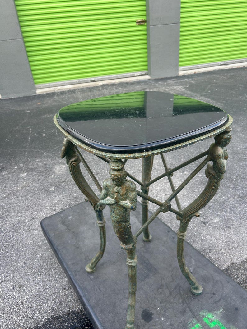 K&ouml;nnte beinhalten: Ein Beistelltisch mit einer schwarzen Marmorplatte und einem gr&uuml;nen Metallgestell mit dekorativen Figuren. Der Tisch hat eine quadratische Platte und drei Beine.