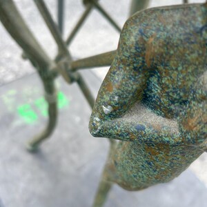 K&ouml;nnte beinhalten: Eine gr&uuml;ne Bronzeskulptur einer stehenden Person mit verschr&auml;nkten Armen. Die Skulptur hat eine strukturierte Oberfl&auml;che und steht auf einem grauen Hintergrund.