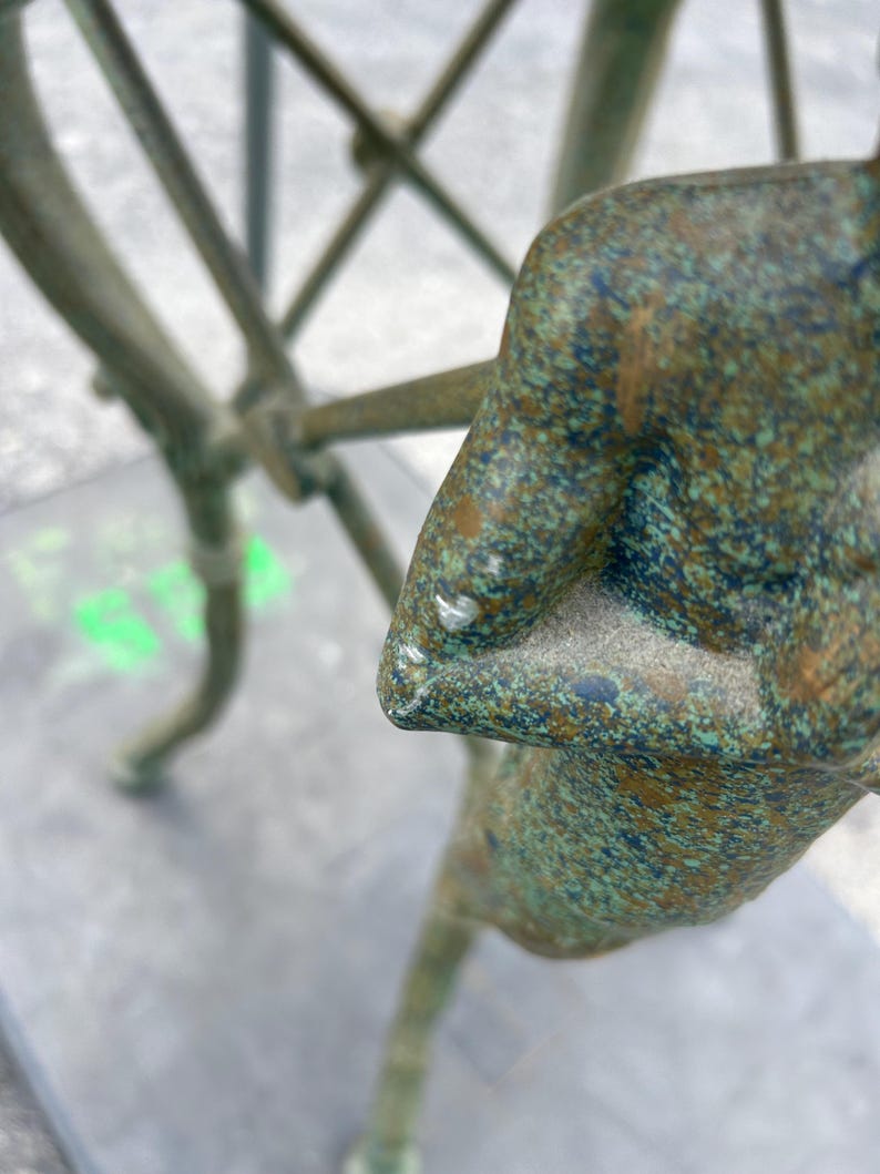 K&ouml;nnte beinhalten: Eine gr&uuml;ne Bronzeskulptur einer stehenden Person mit verschr&auml;nkten Armen. Die Skulptur hat eine strukturierte Oberfl&auml;che und steht auf einem grauen Hintergrund.