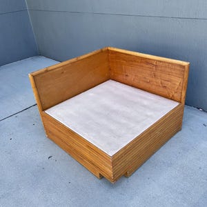 May include: A corner wooden chair with a light gray seat. The chair is made of light brown wood and has a simple, boxy design. The chair is approximately 30 inches wide and 30 inches deep.